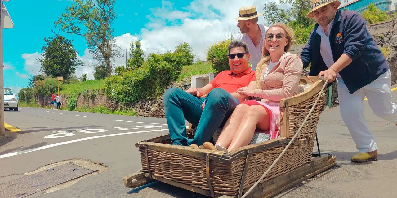 Two guests laughing in a wicker basket sledge on a Monte road, pushed by two men in white clothes and straw hats