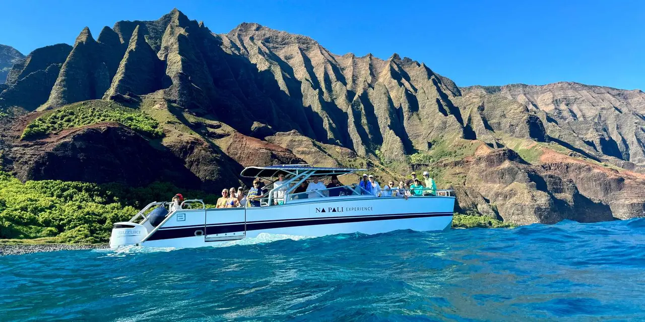 A white power catamaran with passengers on deck cruising in front of the jagged green Na Pali cliffs