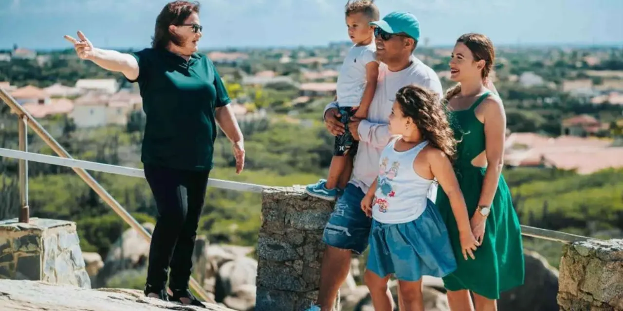A guide pointing out the view to a family of four at a railed overlook above green hills and red-roofed houses
