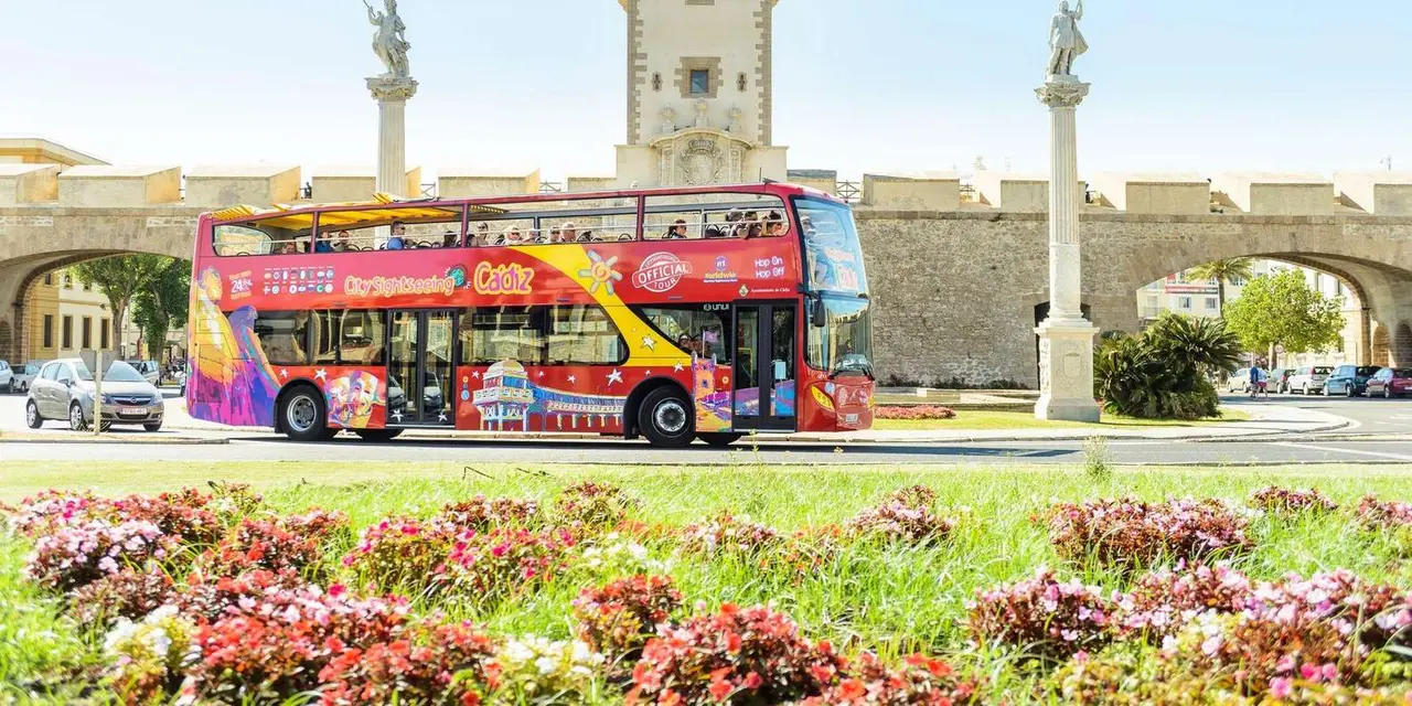 A red open-top double-decker sightseeing bus driving past bright flower beds with stone monuments behind it