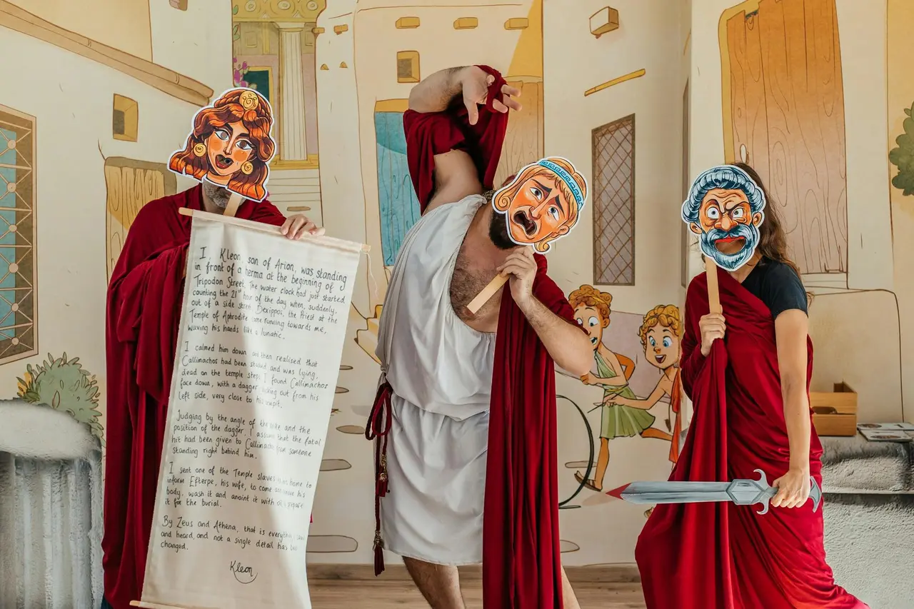 Three performers in red and white robes holding cartoon Greek theatre masks, a scroll and a toy sword in front of a painted ancient street
