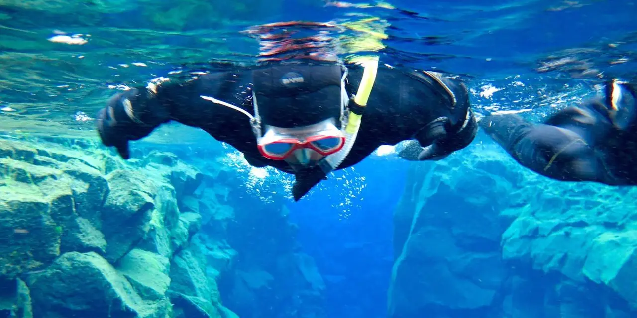A snorkeler in a wetsuit and red-framed mask swimming over rock in clear blue water