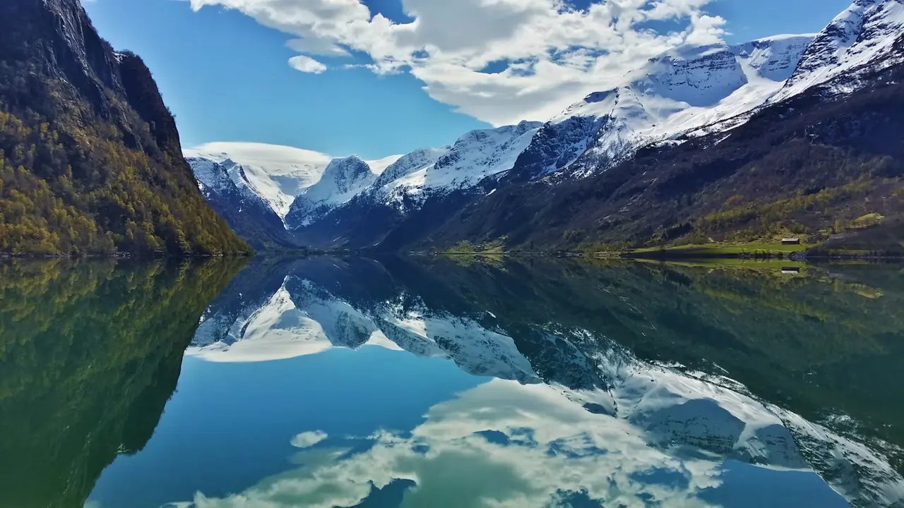 A mirror-calm fjord at Olden reflecting snow-covered mountains and a blue sky with scattered clouds, green hillsides on either side