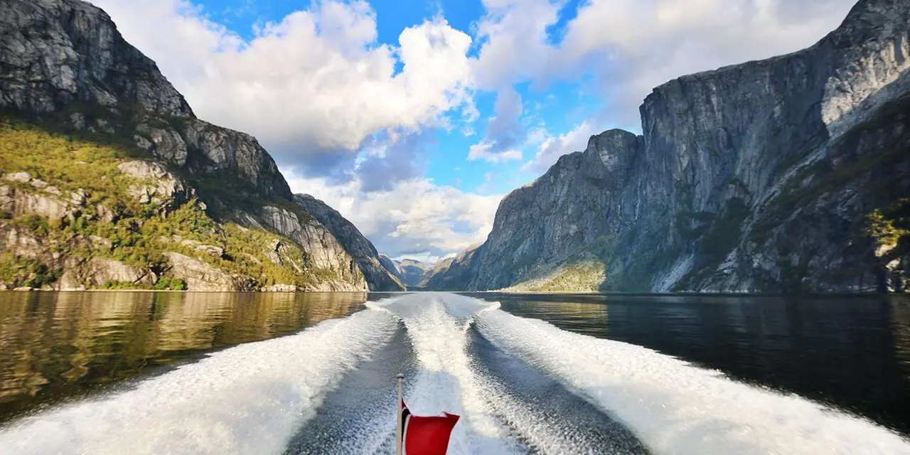 A boat's wake stretching back along a fjord between steep cliffs under a blue sky with clouds