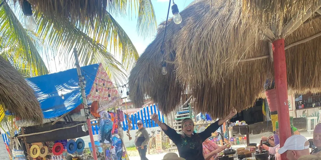 An instructor with both arms raised leading a group seated at tables of stone mortars under thatched palapa roofs on a Mahahual street