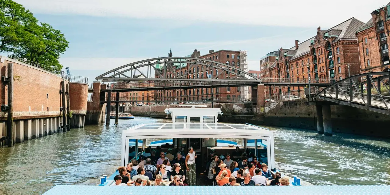 A harbor tour boat full of passengers passing under a steel arch bridge between red-brick Speicherstadt warehouses
