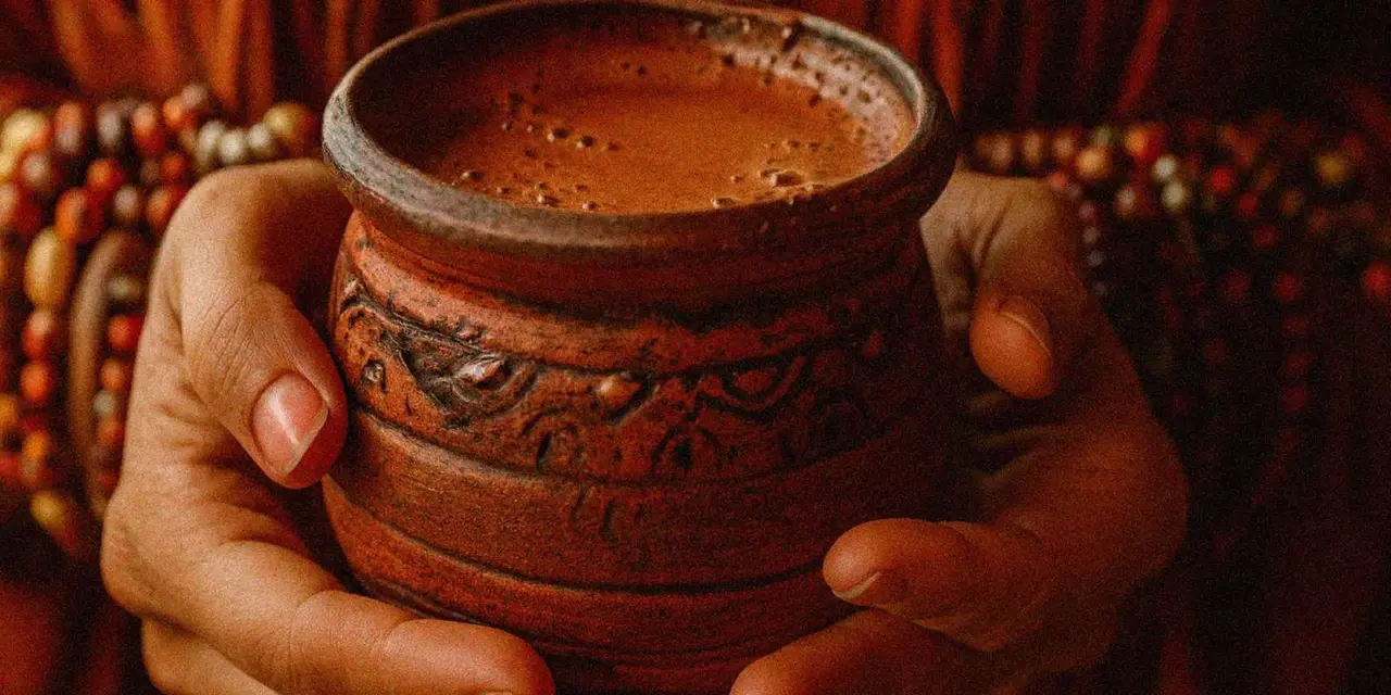 A hand holding a carved clay cup of frothy cocoa beside strands of beads