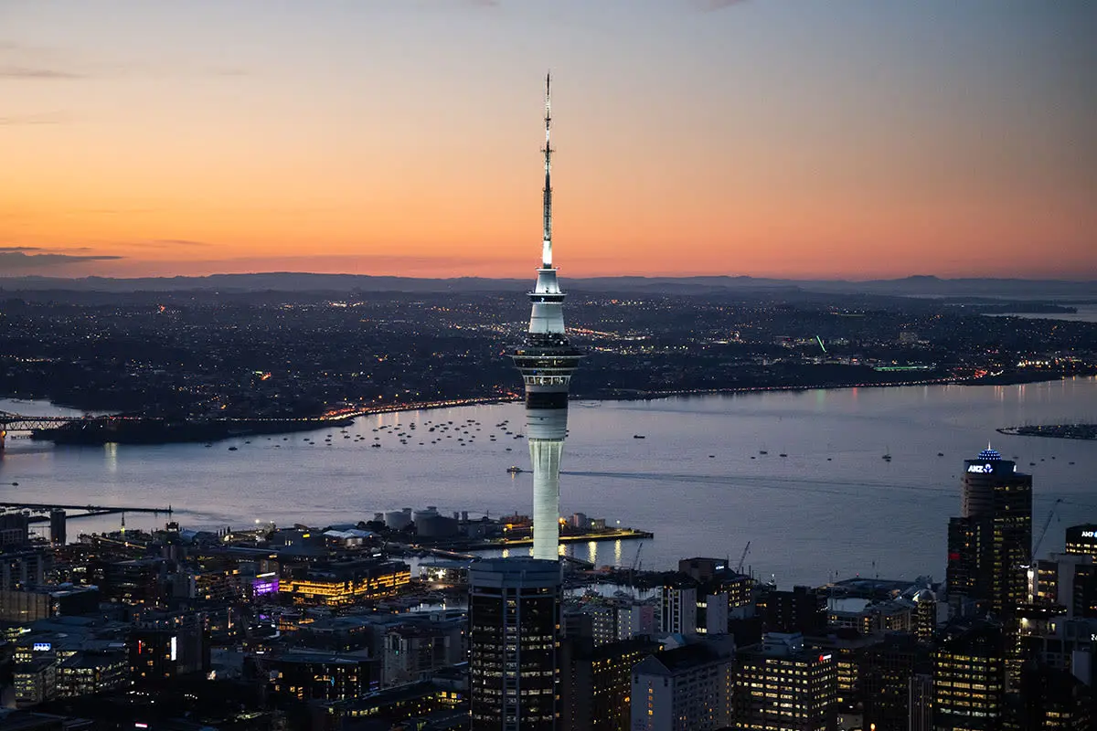 The Sky Tower lit up at dusk above Auckland's city lights, with the harbour and an orange sky beyond
