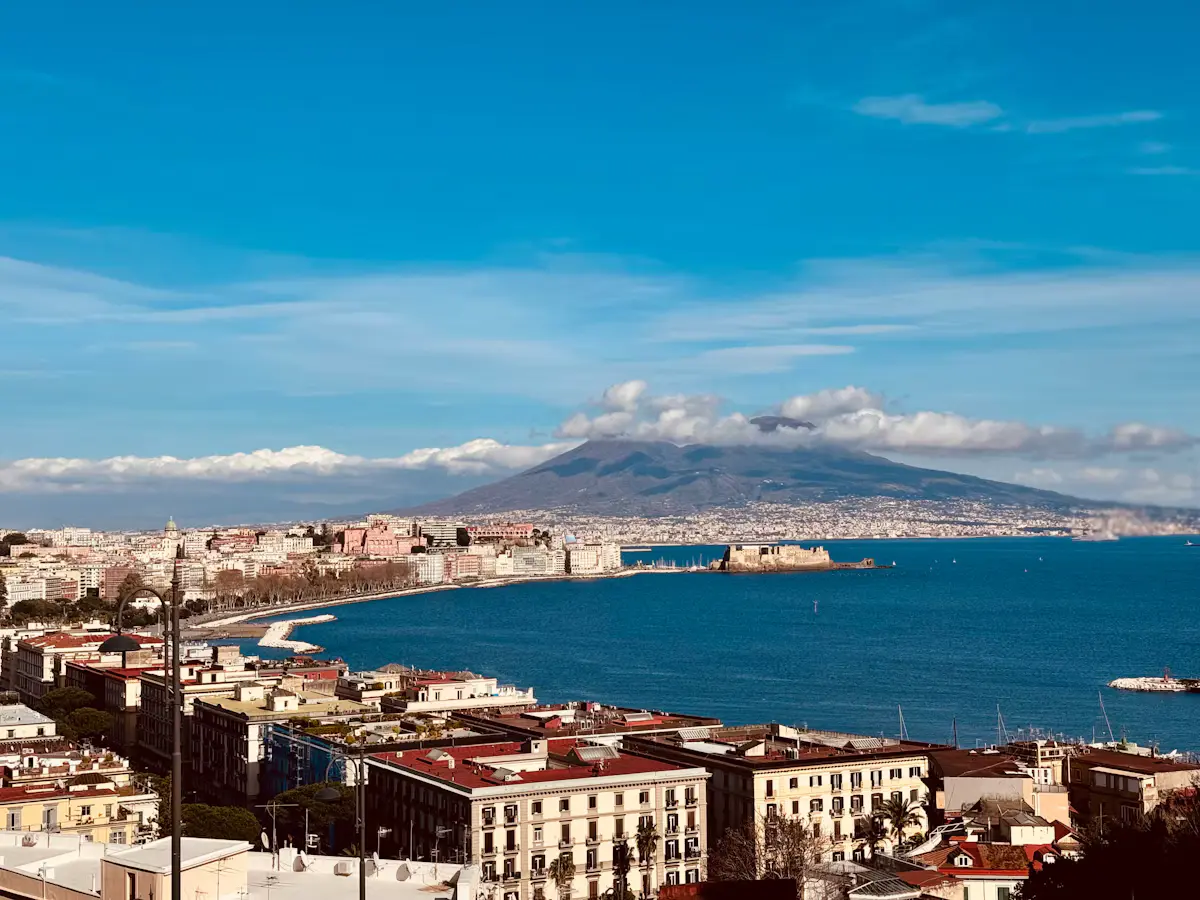 The Bay of Naples from the Posillipo hillside: the curving Chiaia seafront leads out to Castel dell'Ovo on its little island, with Mount Vesuvius rising behind under a blue sky and a band of white cloud