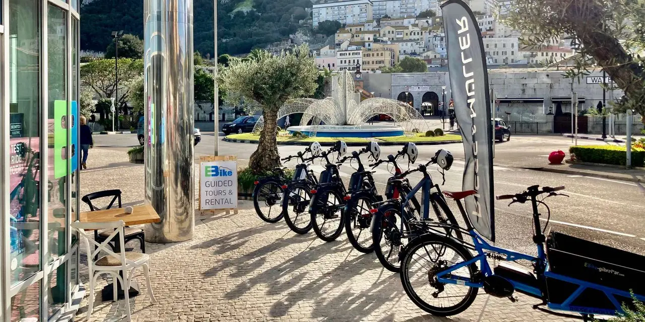 A row of electric bikes parked outside a shop in Ocean Village, with the Rock and hillside houses behind