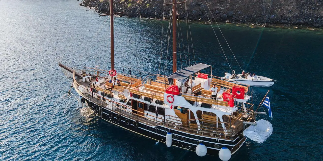 Aerial view of a two-masted wooden boat with benches on its decks, on dark blue water beside a rocky shore