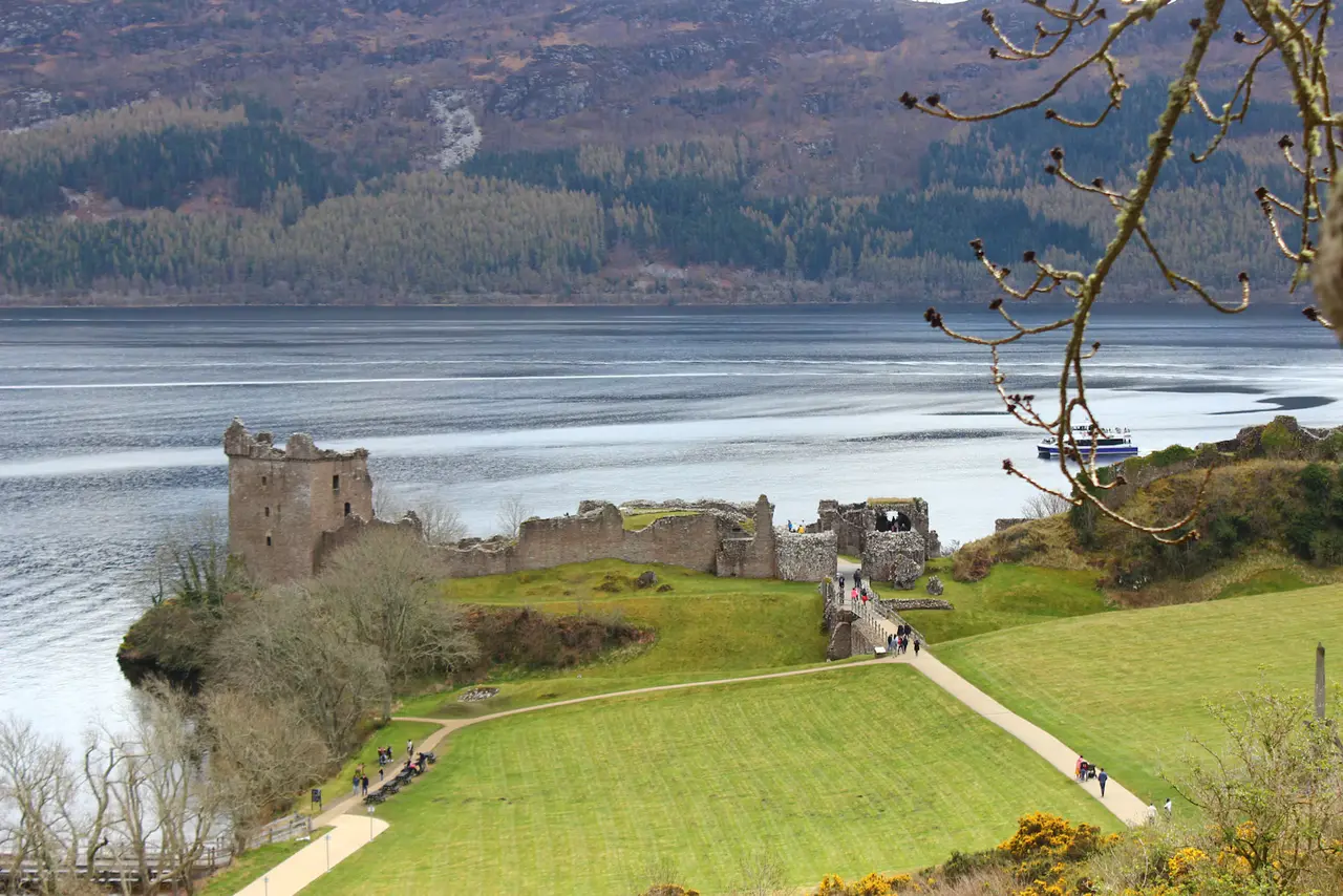Urquhart Castle's ruined tower and walls on a grassy headland above Loch Ness, wooded hills behind and a tour boat on the water