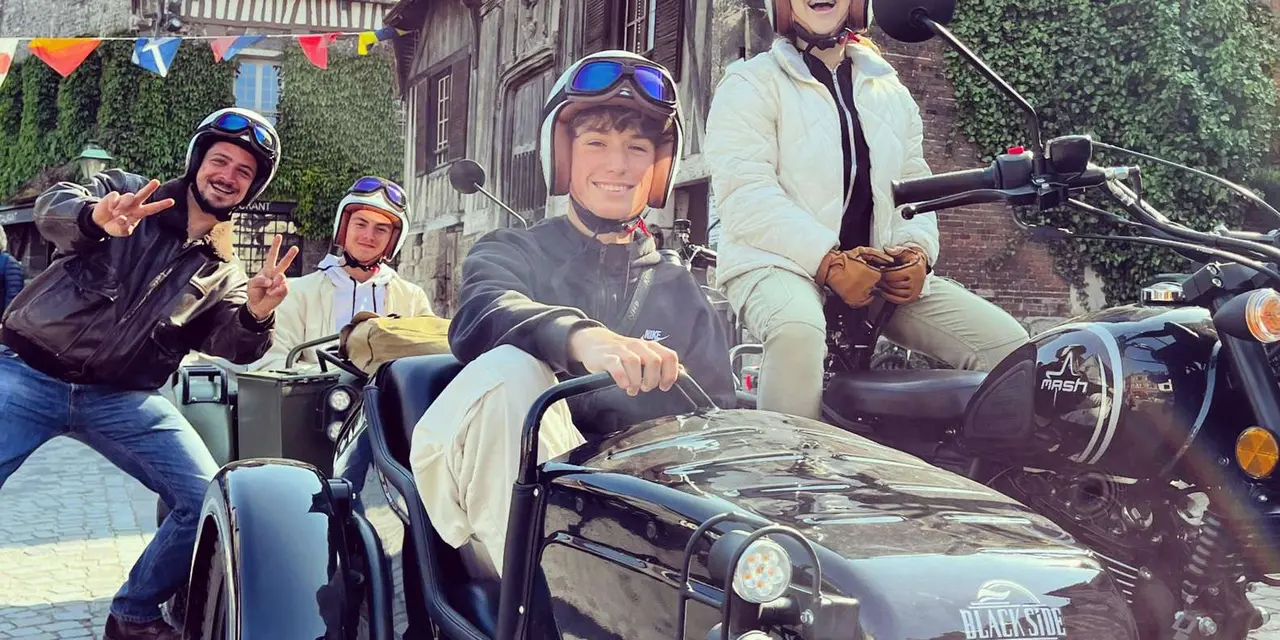 Four people in retro helmets and goggles posing with black sidecar motorbikes on a cobbled street lined with half-timbered buildings