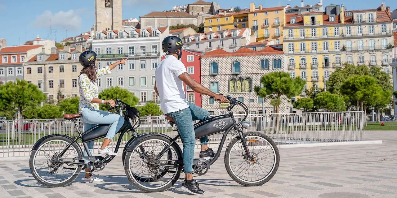 Two people on black electric bikes in helmets, pointing at the pastel apartment blocks and church tower climbing the hill behind a paved waterfront square