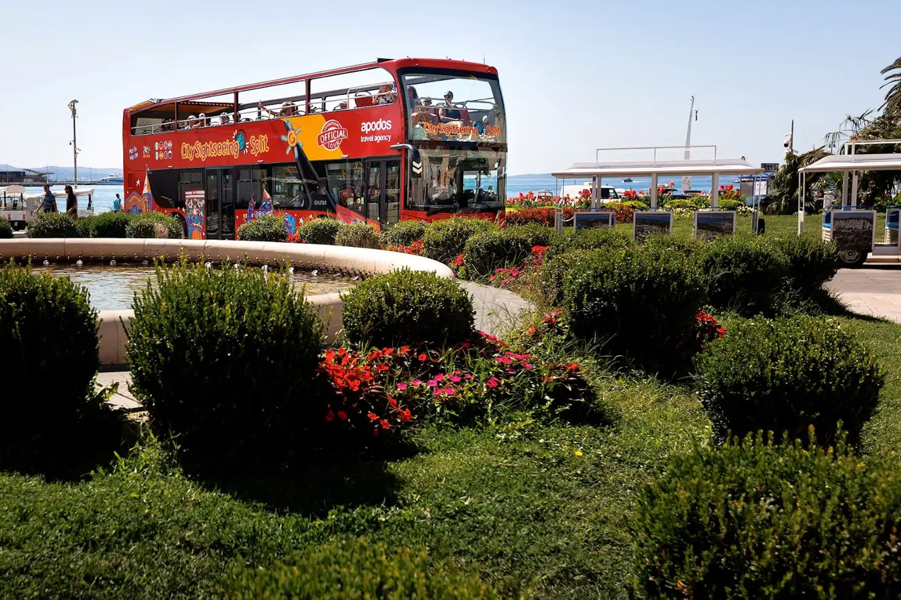 A red open-top sightseeing bus parked behind a fountain and flowerbeds by the sea