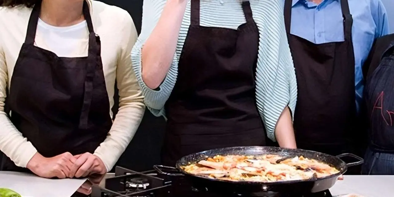 Three people in black aprons tasting from a large pan of paella at a cooking class