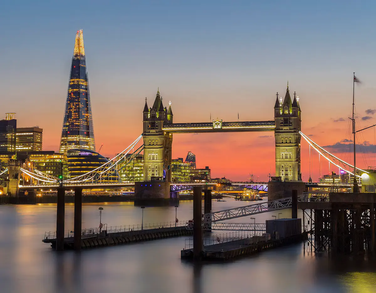 Tower Bridge and the Shard on the Thames at sunset, lit against an orange and pink sky, with a pontoon in the foreground water