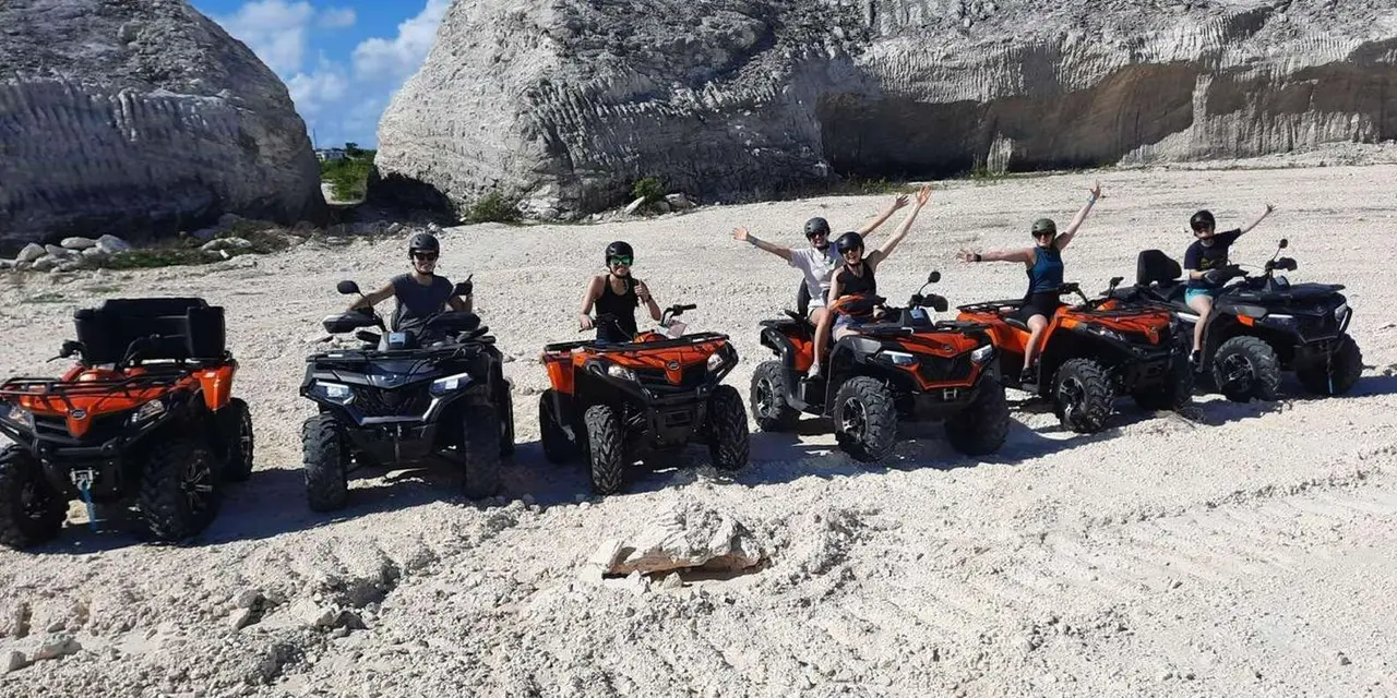 Riders waving from orange and black ATVs lined up on white sand below pale limestone cliffs