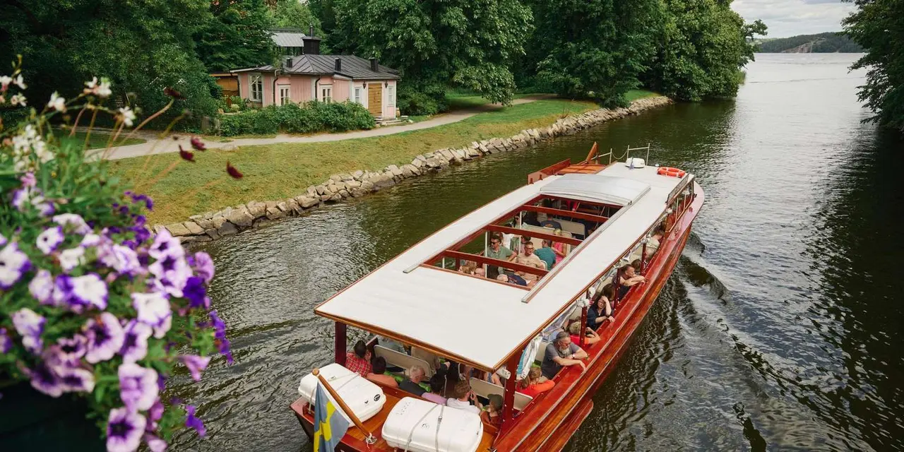 A varnished wooden boat full of passengers on a green canal with trees and flowers along the bank