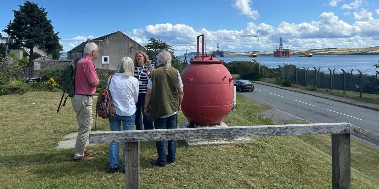 A guide talking to a small group beside a red naval mine on a grassy bank above the firth, with oil rigs in the distance