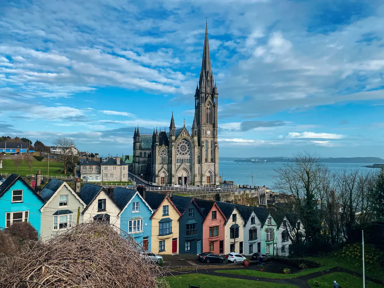 A row of brightly painted houses stepping downhill in front of St Colman's Cathedral in Cobh, with Cork Harbour and a blue sky behind